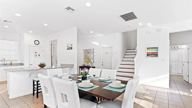 A modern kitchen with white cabinetry, a central island, and a dining area with white chairs and a brown table. - Grand Executive Deluxe 4BR Pool Home Near Disney Parks (Davenport)