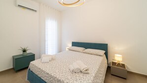A bedroom with a bed, a bedside table with a lamp, a potted plant, and an air conditioning unit.