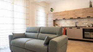 A modern living room with a grey sofa, a kitchen with wooden cabinets, and a potted plant on the counter.