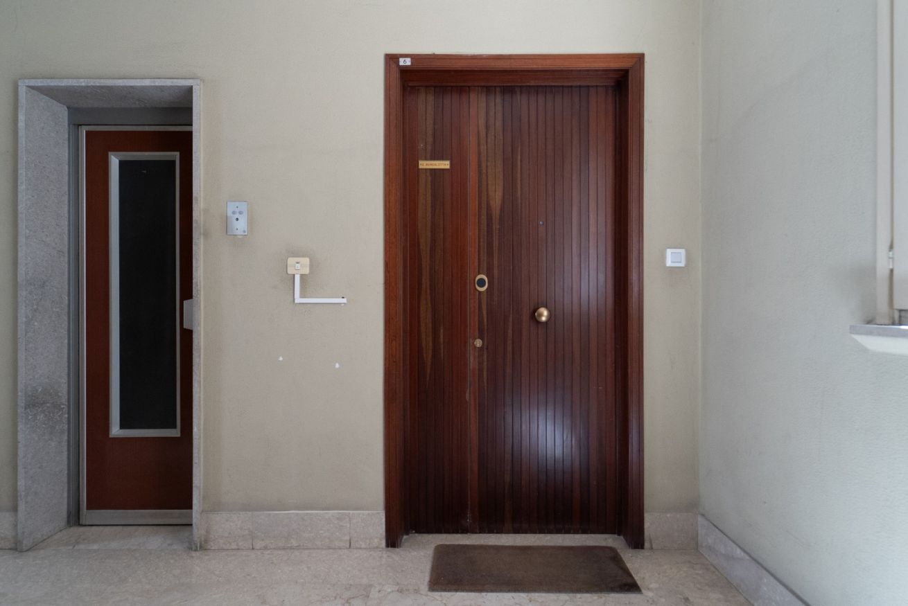 Puerta de madera cerrada con picaporte de latón, felpudo y un interruptor de luz en la pared.