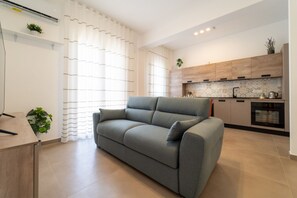 A modern living room with a grey sofa, a wooden dining table, and a kitchen area with wooden cabinets and a tiled backsplash. - GuestHost - Dimora Dei Consoli - Charming Apartment in Sicily (Siracusa)