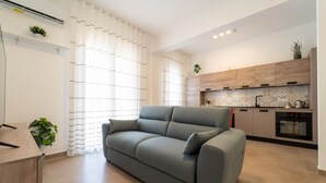 A modern living room with a grey sofa, a wooden dining table, and a kitchen area with wooden cabinets and a tiled backsplash.