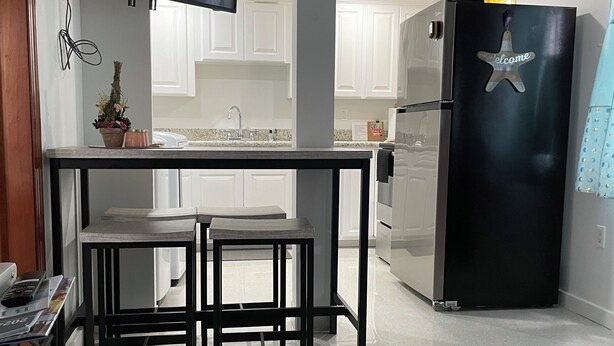 A modern kitchen with white cabinets, a black refrigerator with a 'Welcome' sign, a TV mounted above the counter, and two black stools. - MATUK&ROSE, Cozy , 1  Bedroom , 1 BathroomUnit , complete with AC,WiFi,smart TV
 (Belize City)