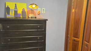 A dark wooden dresser with multiple drawers against a light purple wall, a small table lamp, and a painting of bottles. - MATUK&ROSE, Cozy , 1  Bedroom , 1 BathroomUnit , complete with AC,WiFi,smart TV
 (Belize City)