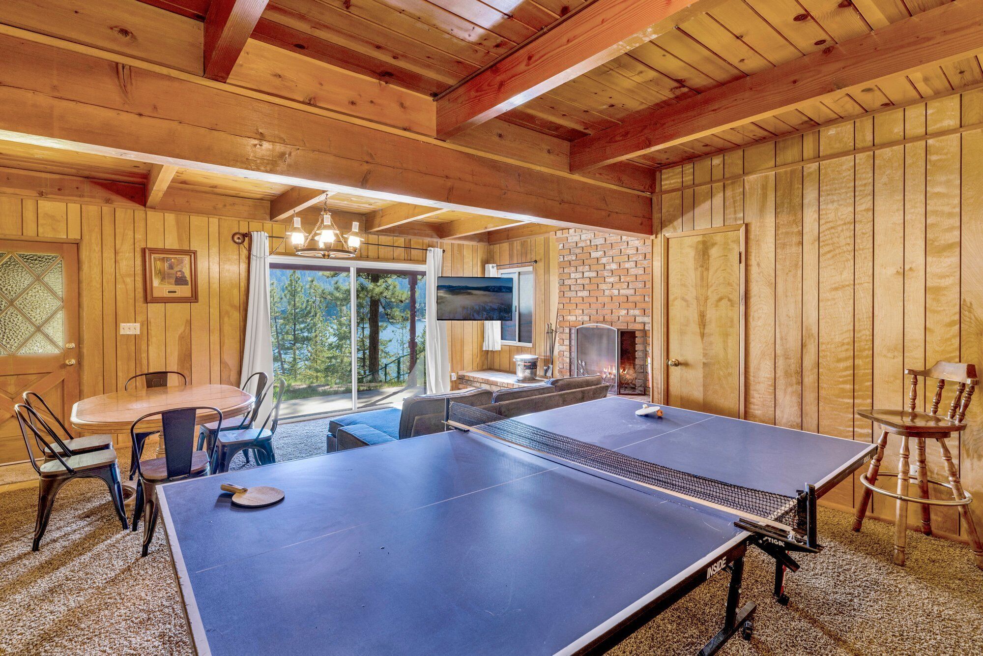 A room with a ping pong table, a fireplace, and a large window with a view of trees.