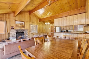 A cozy cabin interior with a stone fireplace, wooden dining table, and a kitchen area. - Inspiration Point Retreat #2470 (Big Bear Lake)