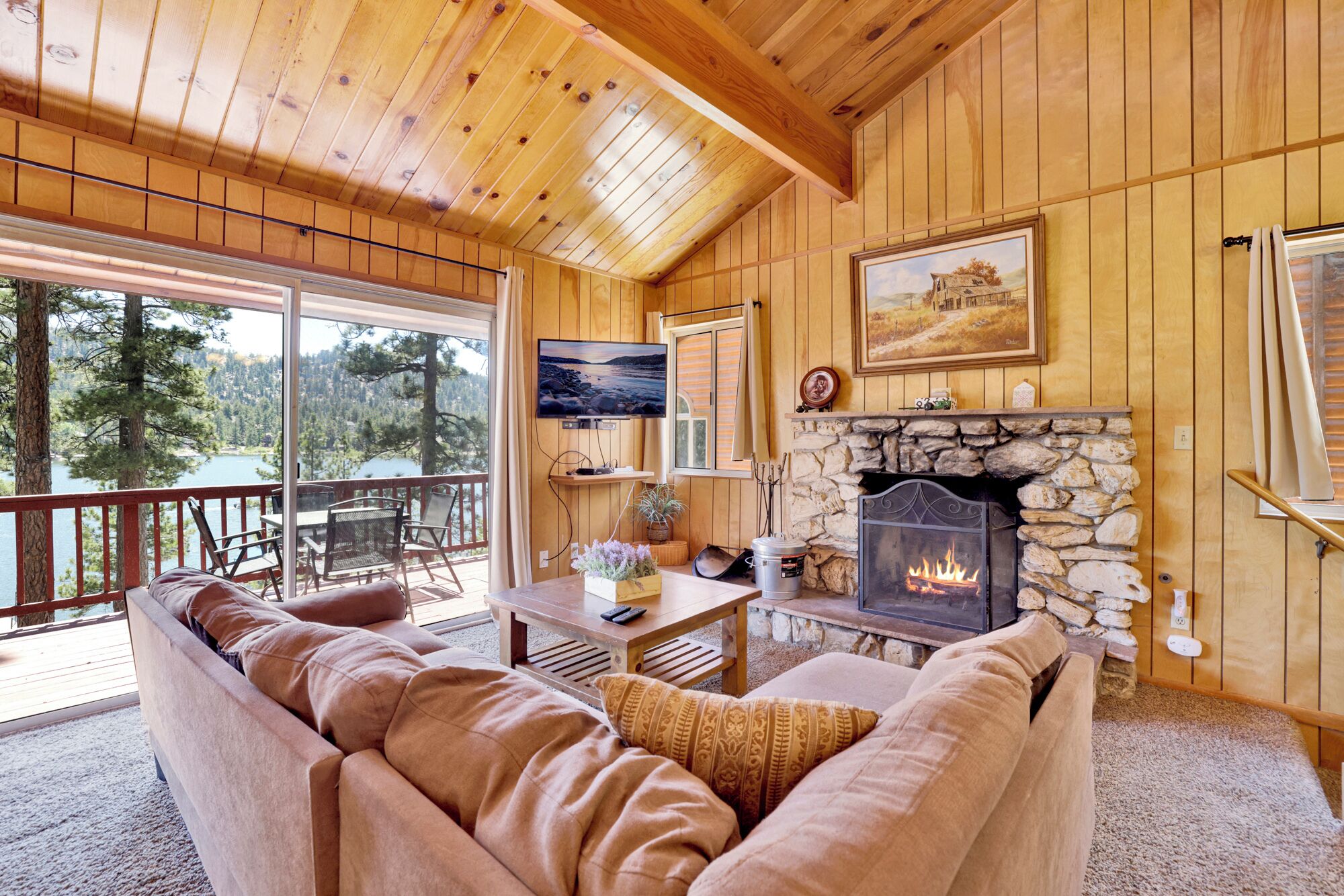 L’intérieur chaleureux d’un chalet avec une cheminée en pierre, des murs en bois, une grande fenêtre donnant sur une forêt et un coin salon confortable.