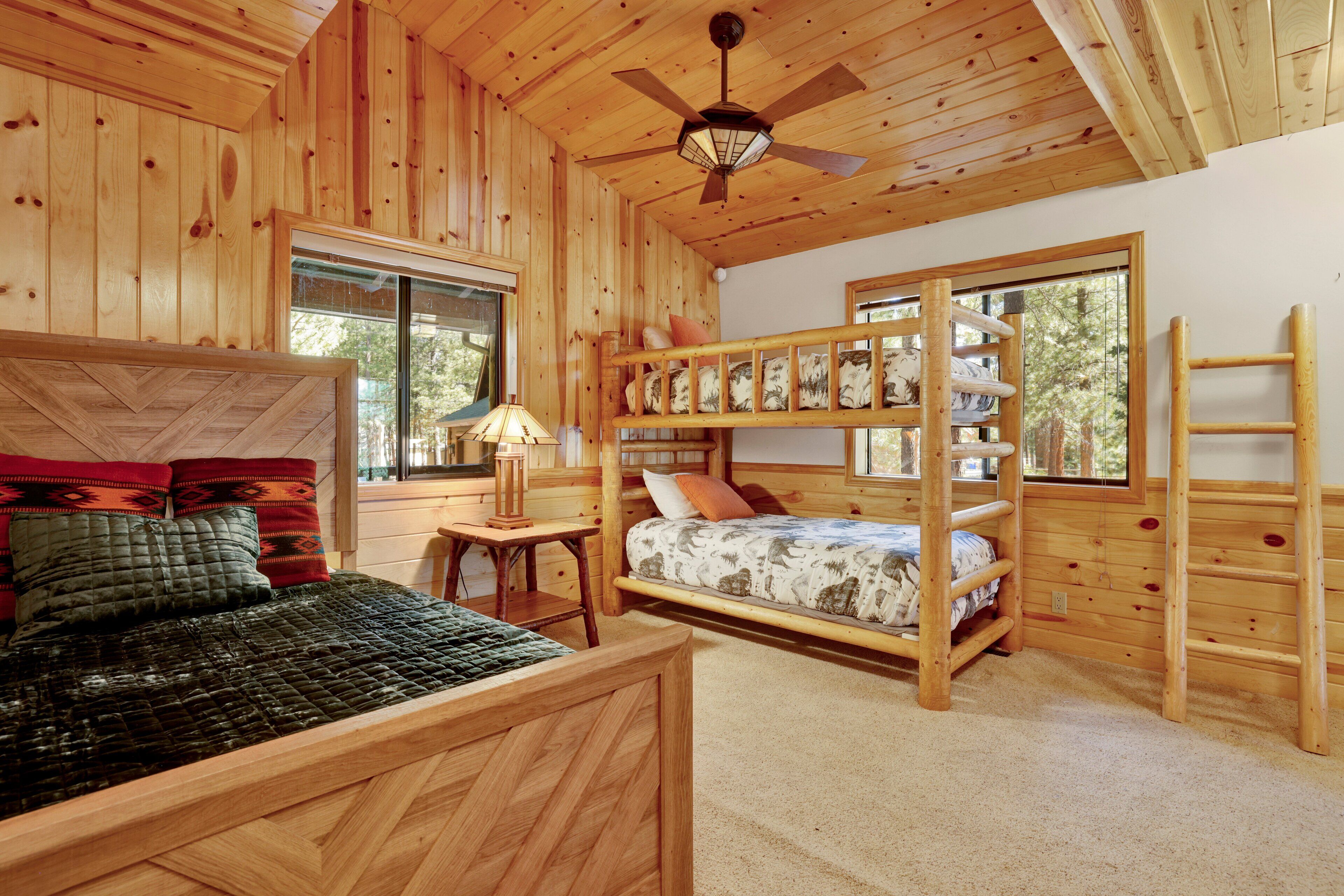 A wooden cabin bedroom with a bunk bed, a single bed, a ceiling fan, and a window providing a view of the outdoors.