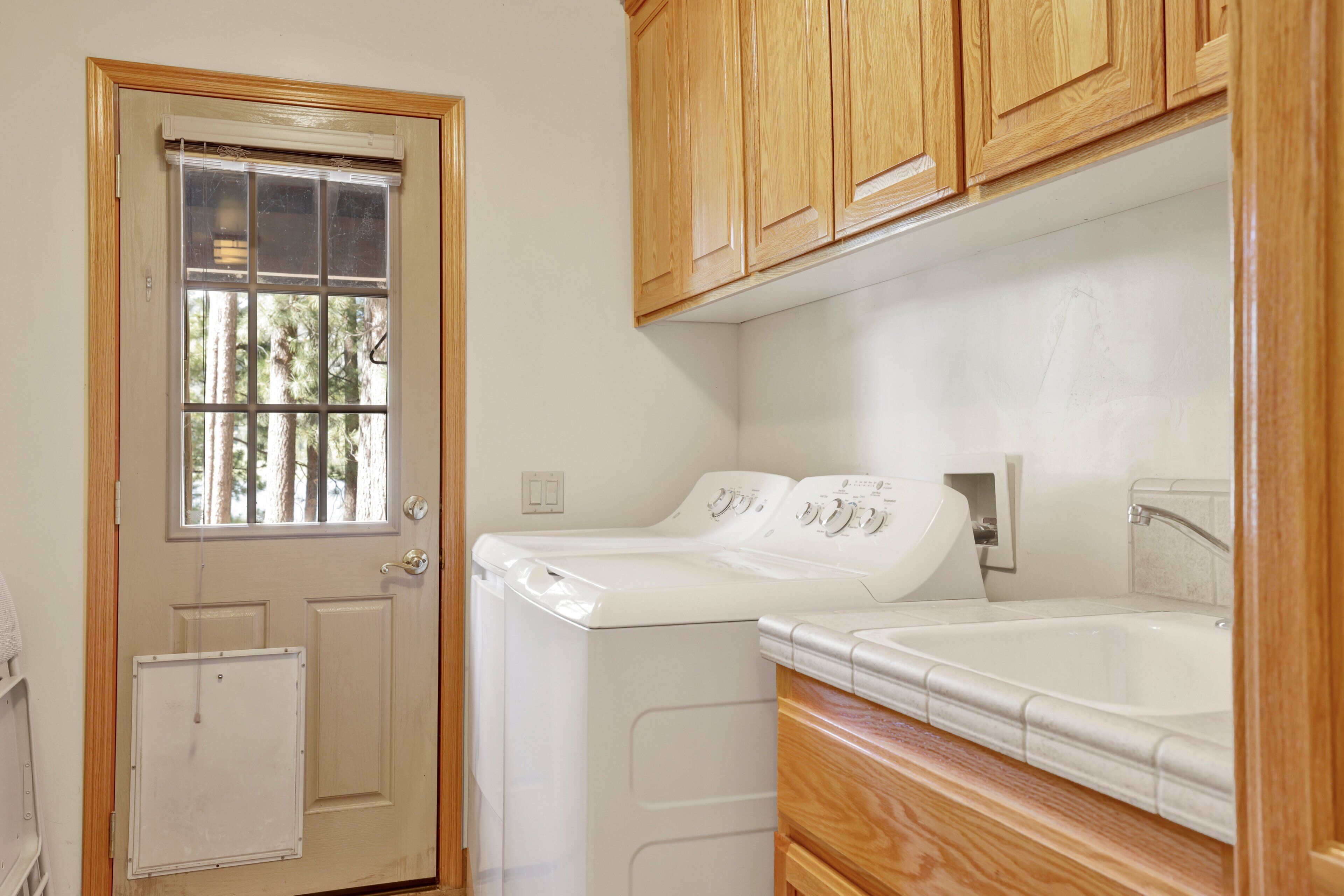 A laundry room with a washer and dryer, wooden cabinets, and a window with a view of trees.