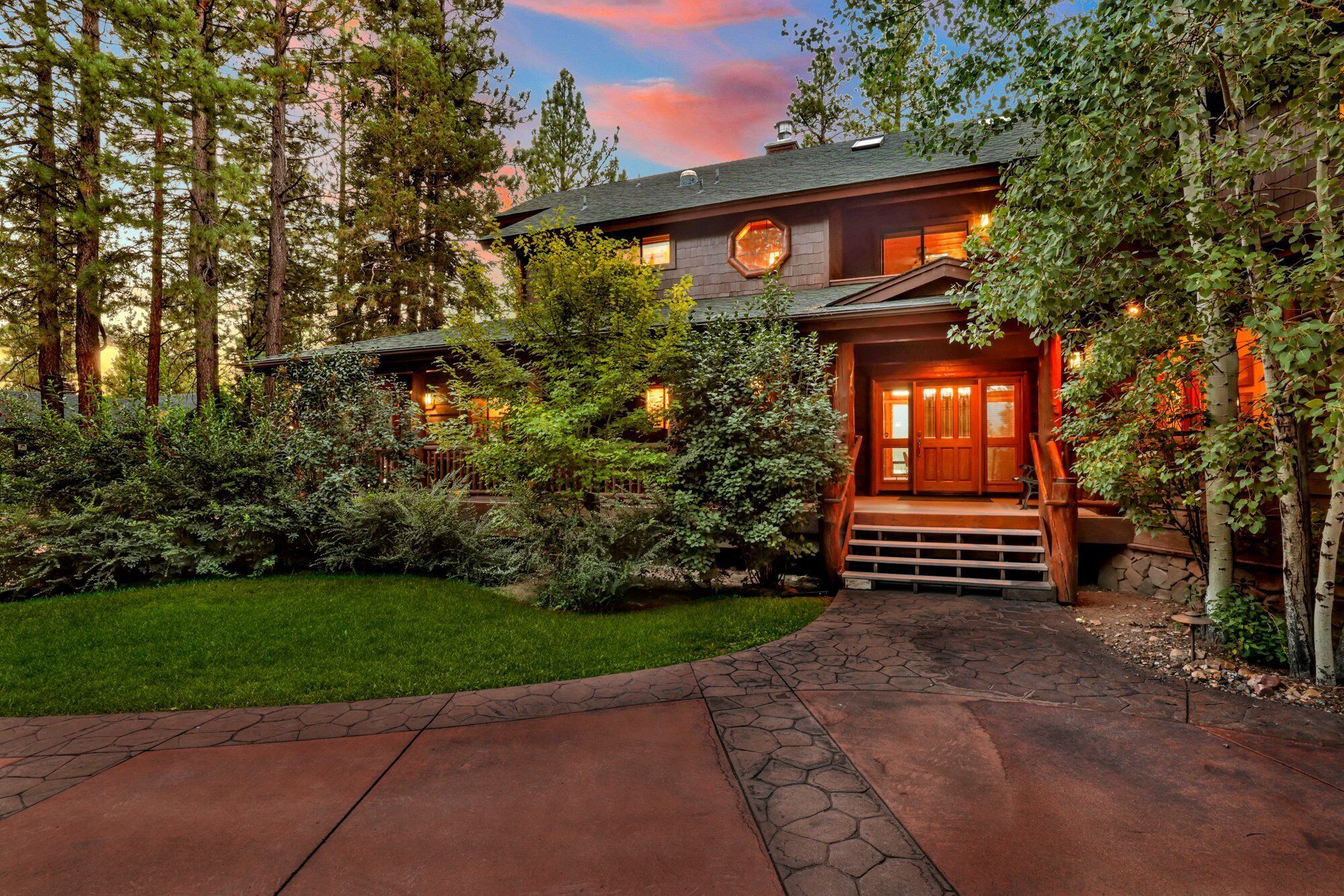 A log cabin with a stone pathway leading to a wooden front door, surrounded by dense greenery and tall trees.