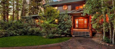 A log cabin with a stone pathway leading to a wooden front door, surrounded by dense greenery and tall trees.