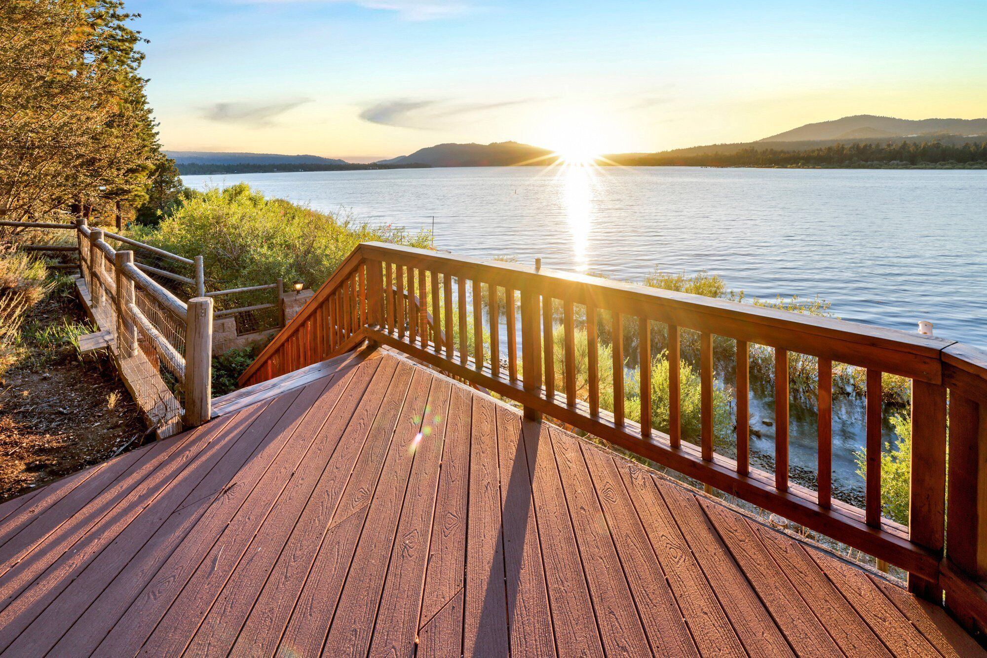 A wooden deck with a railing overlooking a lake at sunset.