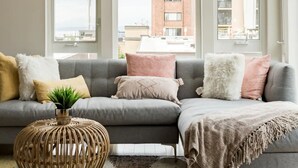 A modern living room with a grey sectional sofa, a woven side table, and a view of a cityscape through large windows. - Walk to Space Needle from a Cap Hill Loft w/VIEWS (Seattle)