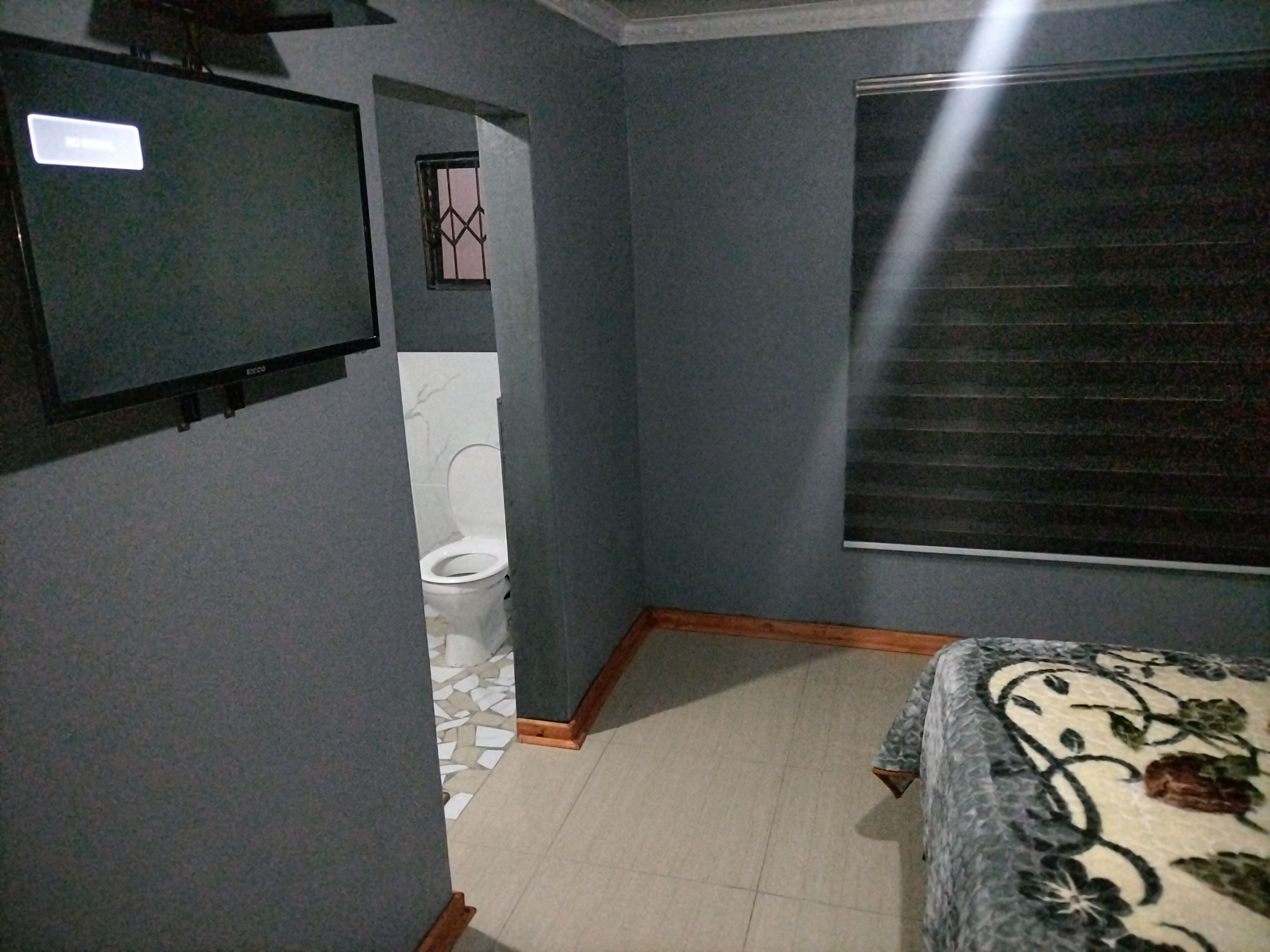 A bedroom with a bed, a television mounted on the wall, a bathroom with a toilet, and a window with blinds.