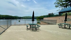 Une terrasse en béton, dotée de tables de pique-nique et de parasols, surplombe un plan d’eau et est délimitée par une barrière métallique.