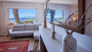 Un moderno salón con un sofá gris, un tapete rojo estampado y un ventanal con vistas al mar y a las montañas.