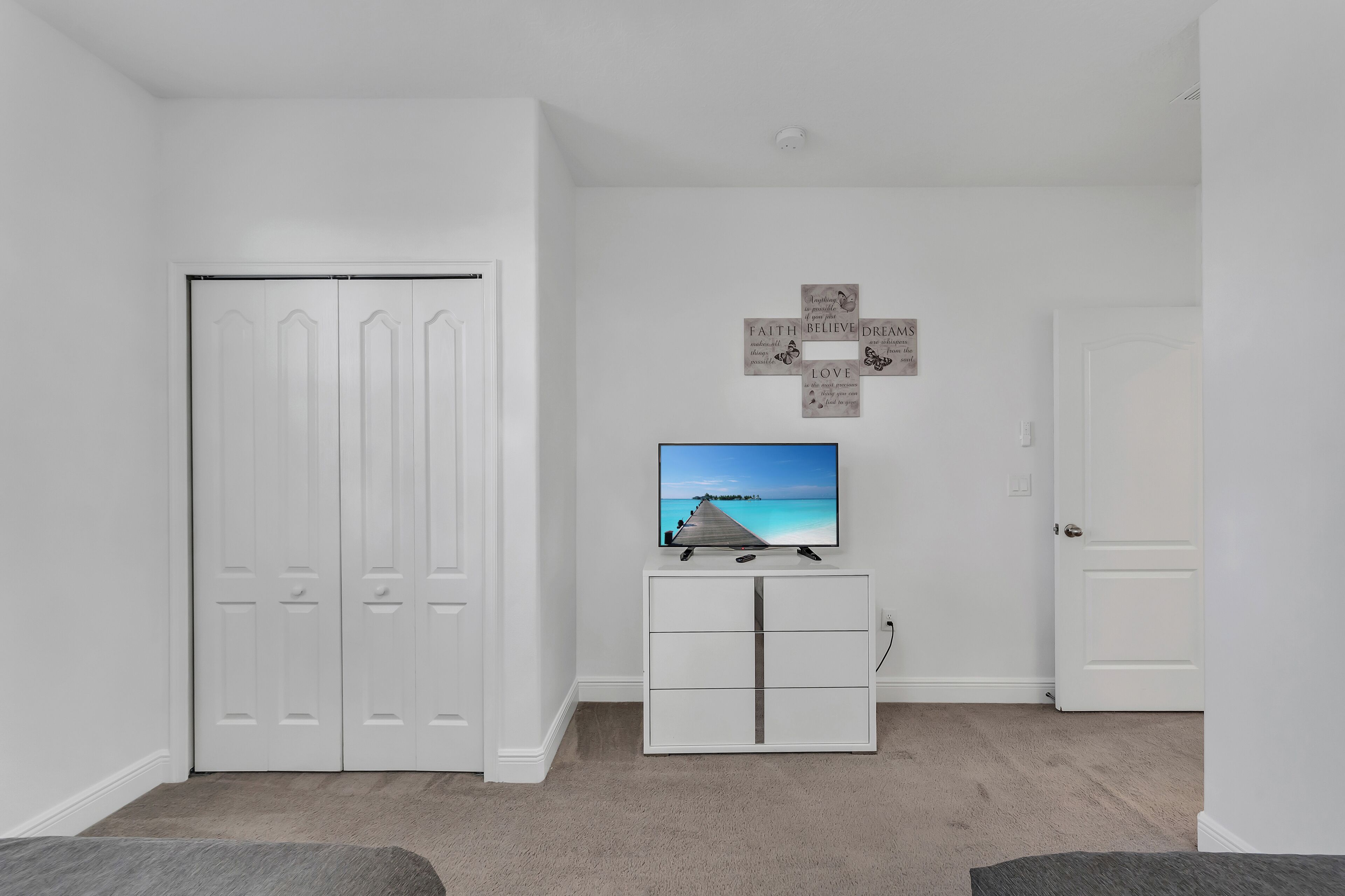 Een kamer met een witte kledingkast, een wit kastje met een tv waarop een strandafbeelding te zien is, en een muur met motiverende citaten.