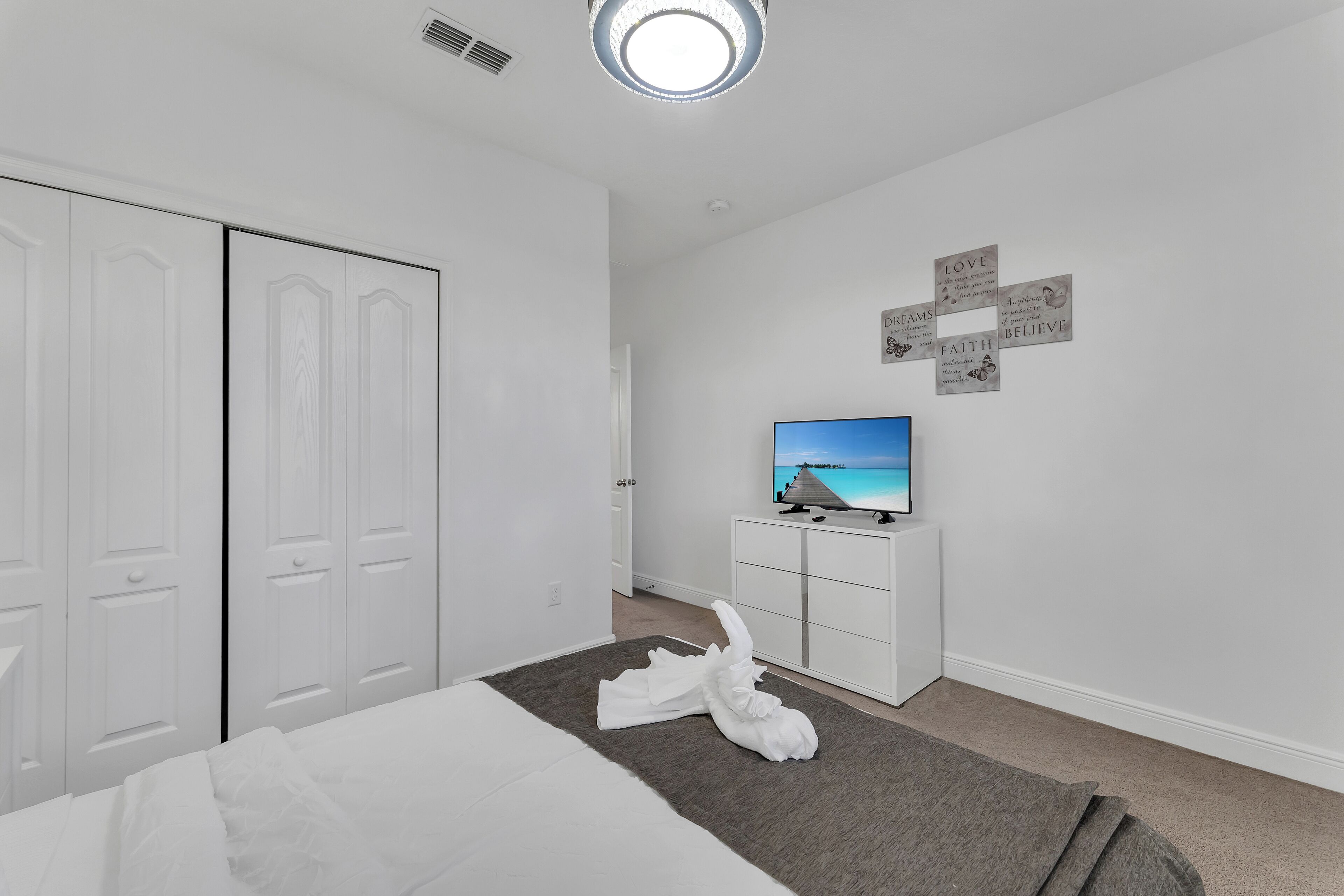Quarto de hotel com cama, televisão de tela plana, guarda-roupa e arte motivacional na parede.