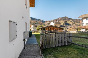 A narrow alleyway leading to a wooden building with a sloped roof, surrounded by a wooden fence and a mountain backdrop.