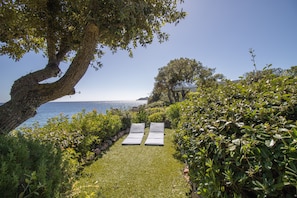 A garden with a pathway leading to a seating area with two chairs, surrounded by greenery and overlooking a body of water.