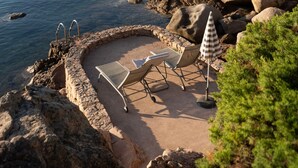 Una playa rocosa con una zona de estar circular, una escalera y una sombrilla.