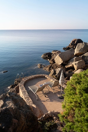 A rocky beach with a circular seating area, a ladder, and a beach umbrella.
