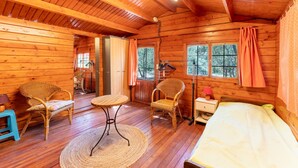Interior de una cabaña de madera con una cama, dos sillas de mimbre, una mesita y una ventana con cortinas.