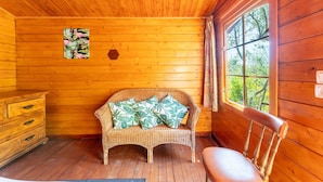Interior de una cabaña de madera con un sofá de mimbre, un aparador de madera y una ventana con vistas a árboles.