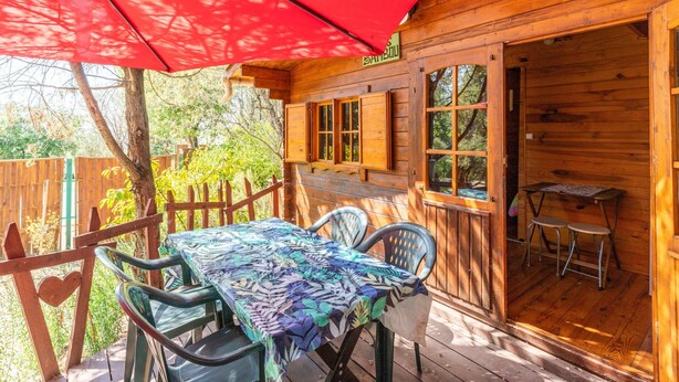 Una terraza de madera con una mesa y sillas, un paraguas rojo y una cabaña de madera con ventanas y una puerta.
