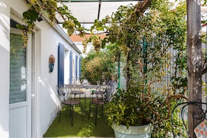 A covered patio with a table and chairs, surrounded by greenery and a white wall.