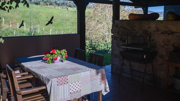 Un espacio exterior cubierto para comer con una mesa lista para dos, un muro de piedra y vistas a un campo verde con aves volando.