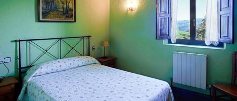 Een slaapkamer met een bed, nachtkastje, lamp, raam met gordijnen, radiator en een schilderij aan de muur.