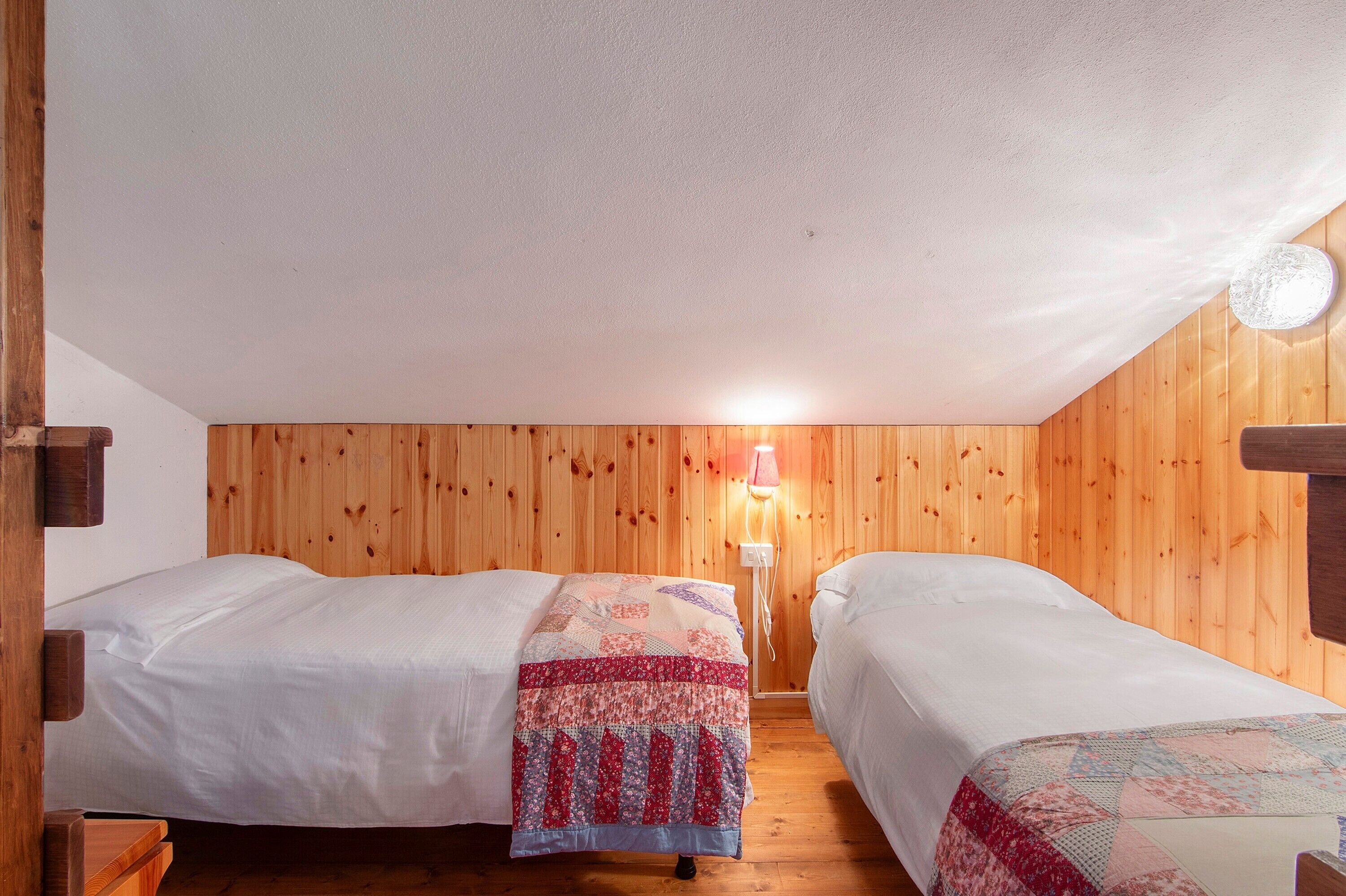 Une chambre avec deux lits, des murs en bois, une lampe fixée au mur et un plancher en bois.