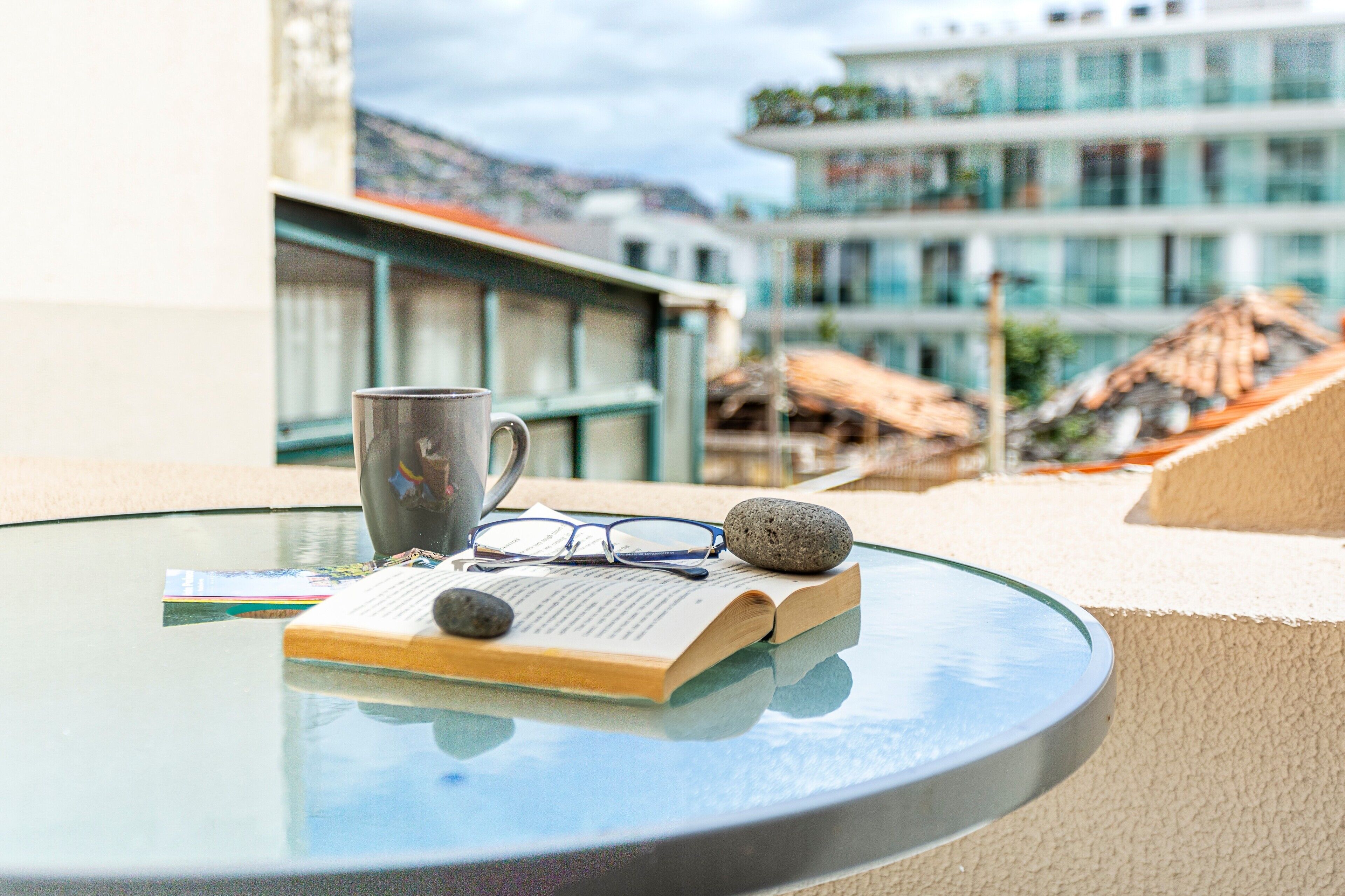 Una mesa de vidrio con un libro, un par de gafas y una taza en un balcón con vistas a edificios y un paisaje montañoso.