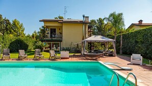 Une piscine avec des chaises longues, un espace de détente couvert et une maison en arrière-plan.