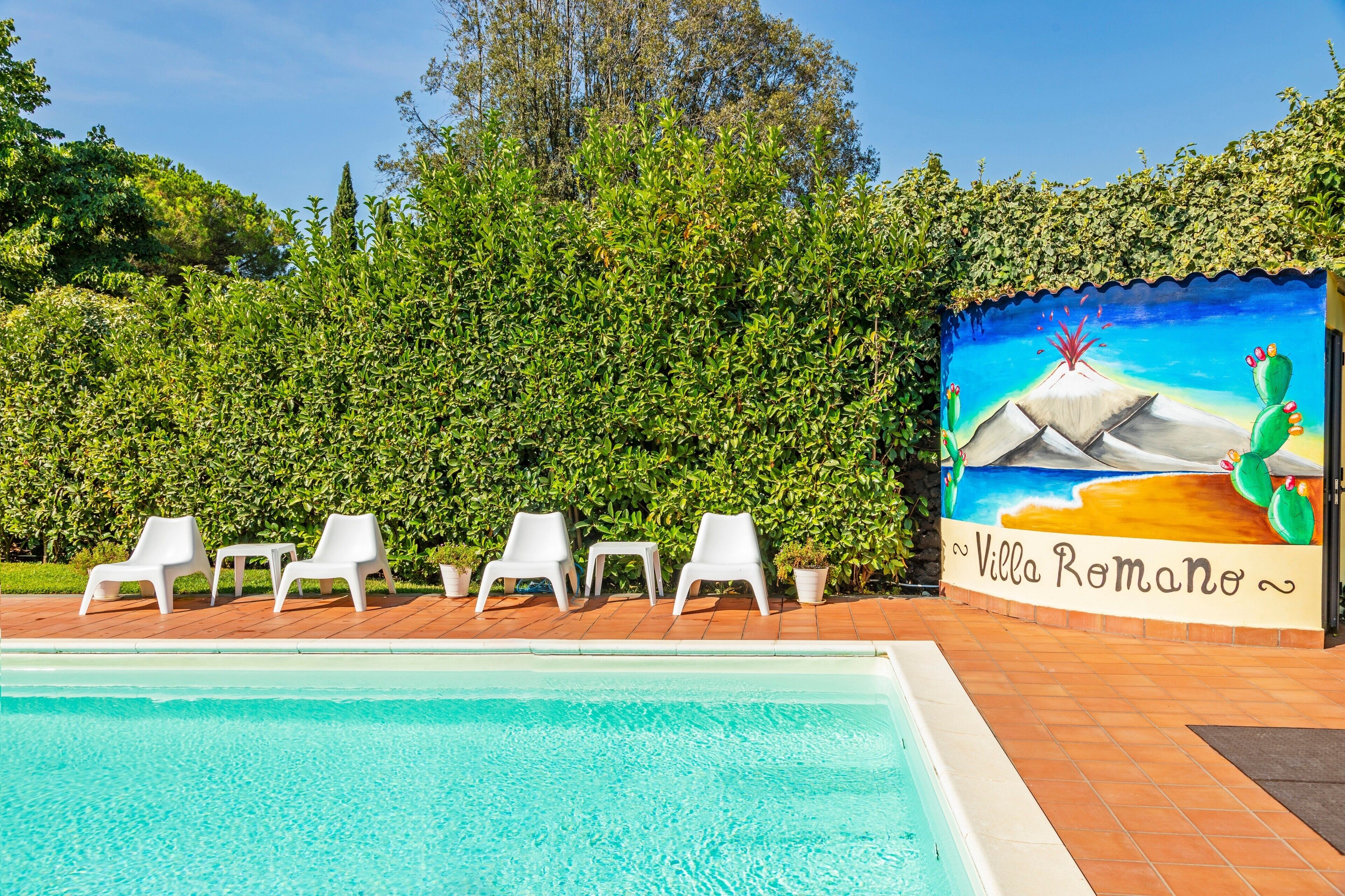 A swimming pool with white lounge chairs, a mural of a beach scene, and a hedge in the background.