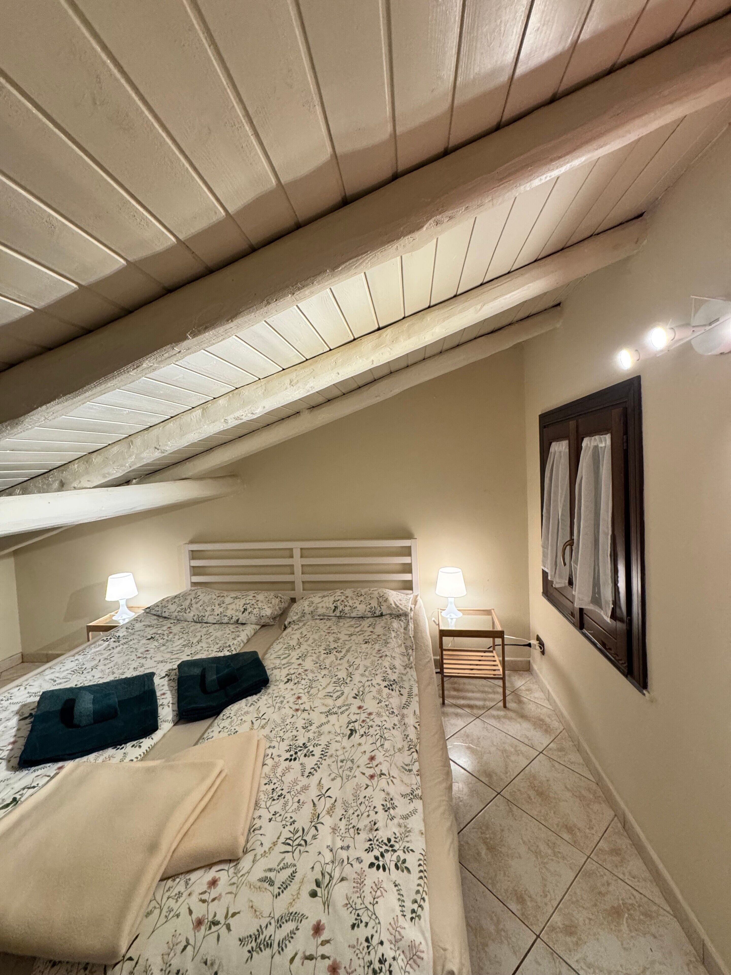 Un dormitorio con techo inclinado, una cama con ropa de cama floral, dos lámparas de noche, una mesita de noche y una ventana con cortinas.