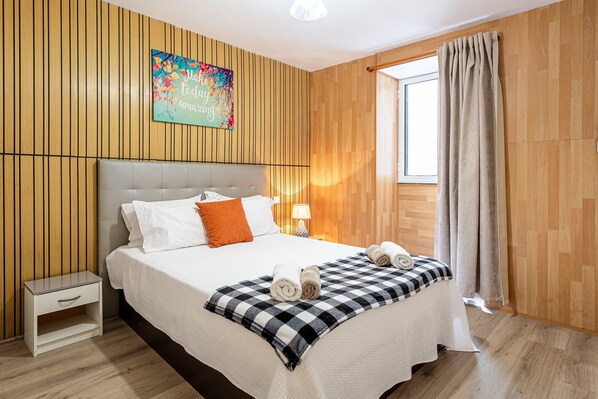 Une chambre d’hôtel avec un mur lambrissé en bois, un lit recouvert d’une couverture à carreaux, une table de chevet et une fenêtre avec des rideaux.