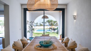 Un coin repas avec une table en bois, des chaises, un vase de fleurs et une vue sur une piscine et des palmiers.