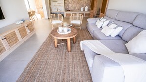 Un salon avec un canapé gris, deux fauteuils en osier, une table basse ronde en bois et un mur en pierre.