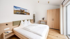 Una habitación de hotel moderna con una cama grande, cabecera de madera, mesita de noche con lámpara, un escritorio pequeño con silla, armario y un ventanal amplio.