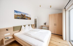 Ein modernes Hotelzimmer mit einem großen Bett, einem hölzernen Kopfteil, einem Nachttisch mit Lampe, einem kleinen Schreibtisch mit Stuhl, einem Kleiderschrank und einem großen Fenster.