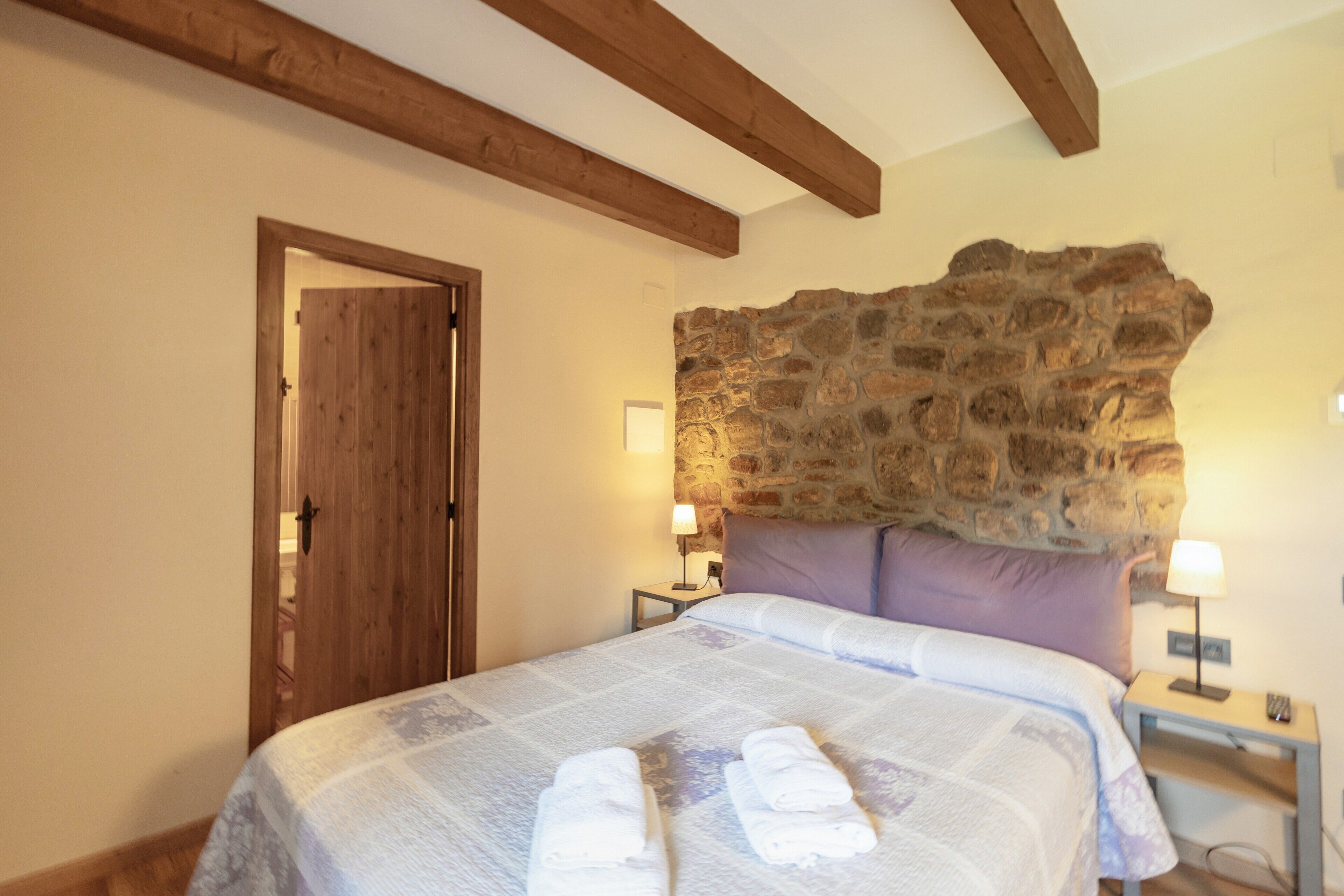 A bedroom with a stone wall, wooden beams, a bed with towels, two bedside tables with lamps, and a door.