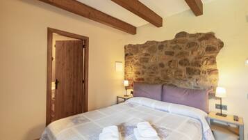 A bedroom with a stone wall, wooden beams, a bed with towels, two bedside tables with lamps, and a door.