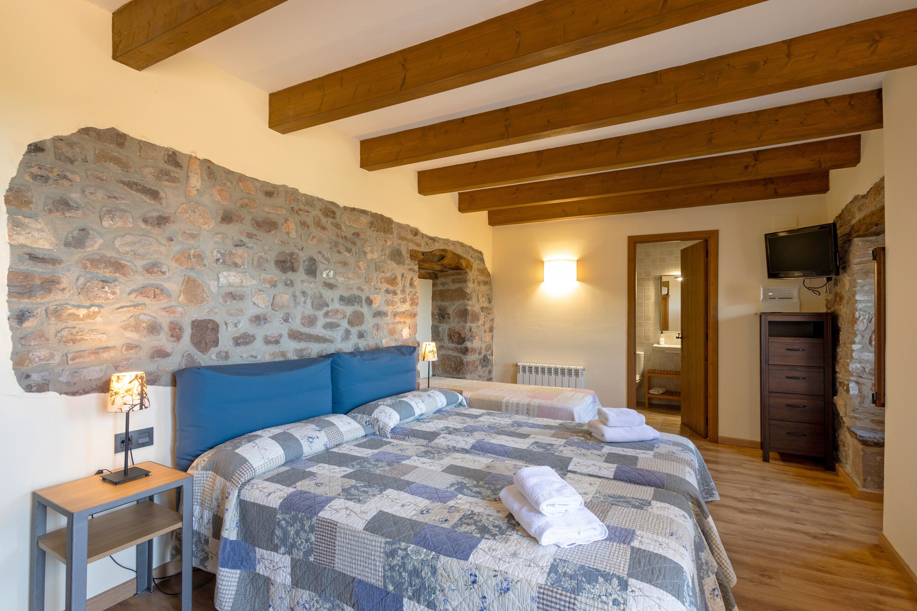 Een slaapkamer met een stenen muur, houten balken, een bed met quilt, nachtkastjes, een televisie en een kledingkast.