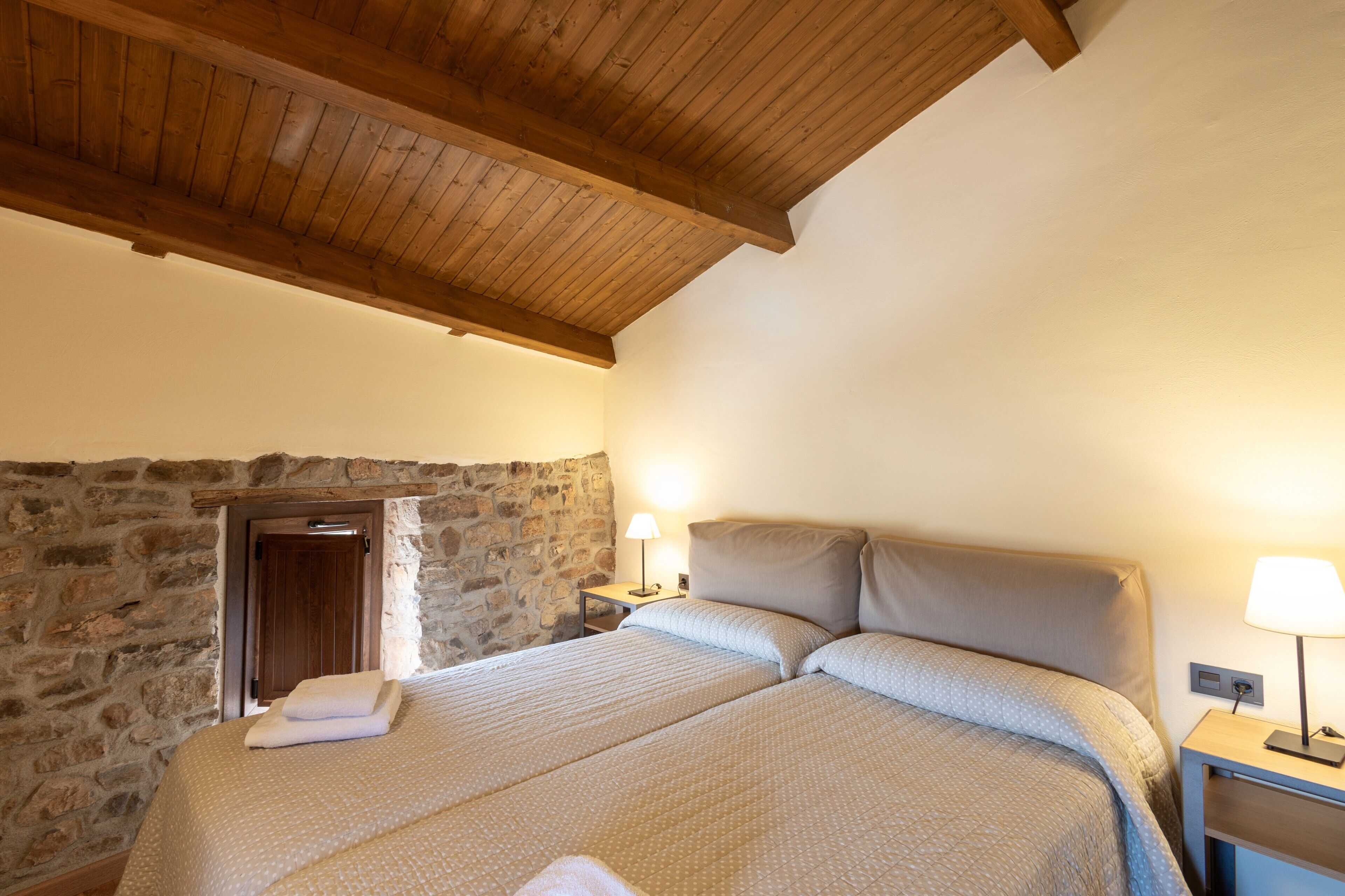 Een slaapkamer met een stenen muur, houten balken in het plafond, een bed met beige beddengoed en bedlampjes.