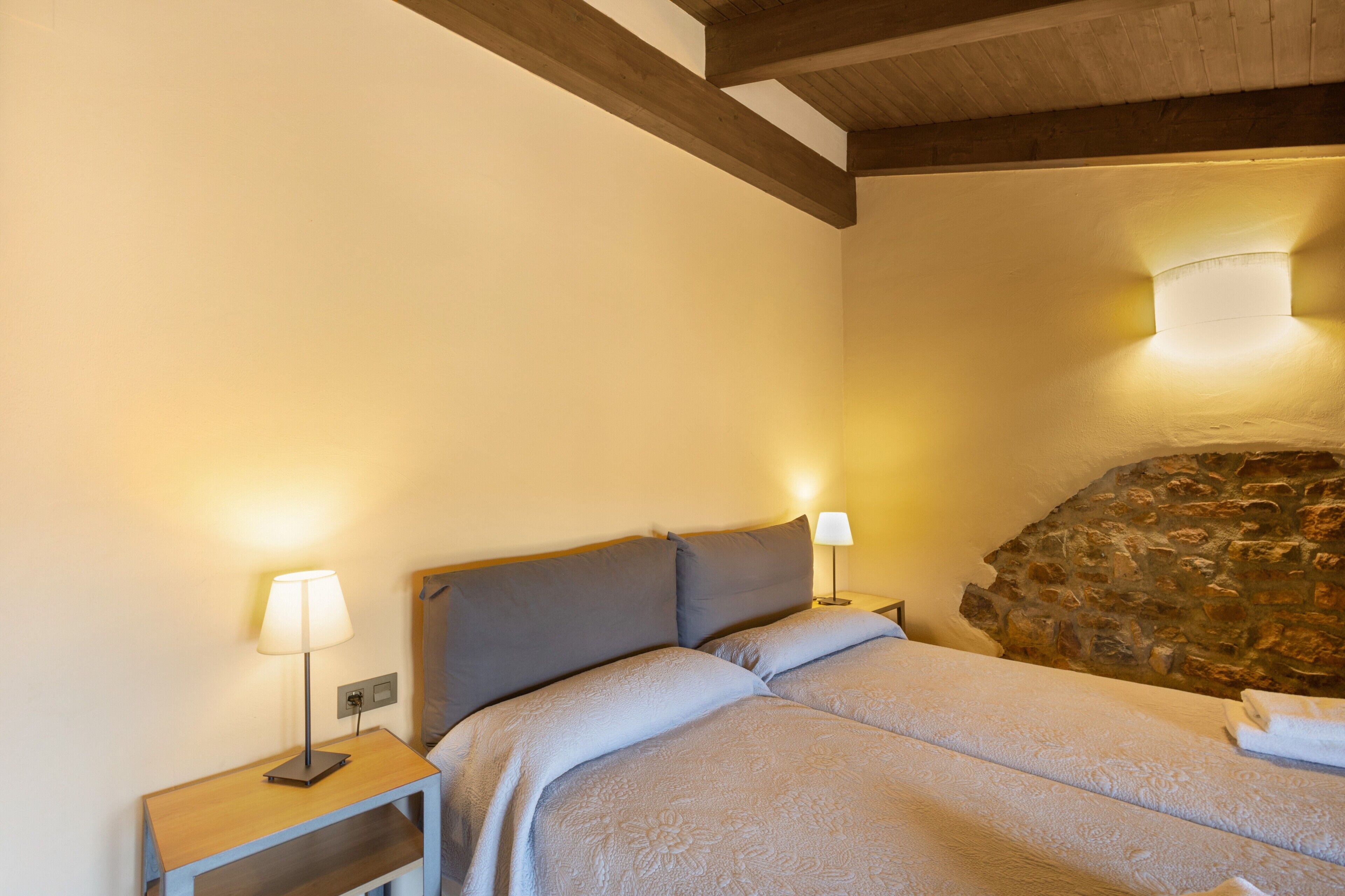 A bedroom with a bed, two bedside tables with lamps, a stone wall, and a wooden ceiling beam.