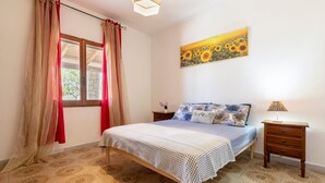 A bedroom with a bed, a nightstand, a lamp, a window with curtains, and a sunflower painting on the wall. - Apartment 'Chef House Sorso' with Shared Terrace and Private Garden (Sorso)