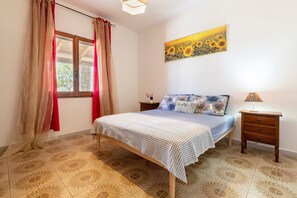 A bedroom with a bed, a nightstand, a lamp, a window with curtains, and a sunflower painting on the wall.
