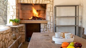 A stone fireplace with a fire, a wooden bench, a table with a fruit basket, and a white tray with cups and bread. - Apartment 'Chef House Sorso' with Shared Terrace and Private Garden (Sorso)